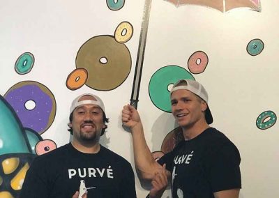 Owners of Purvé Donut Stop Hawaii standing in their first location at the social media wall. There is a unicorn painted on the wall that is pooping donuts like rain